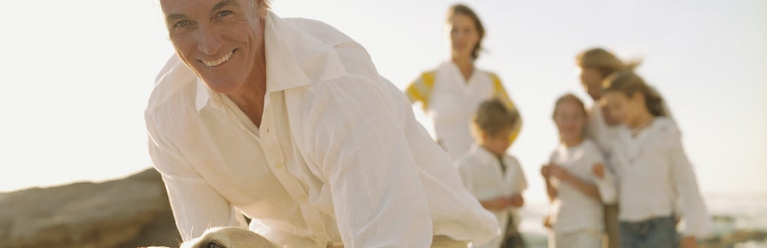 Man smiles while petting a dog on a beach with a woman and four children in the background
